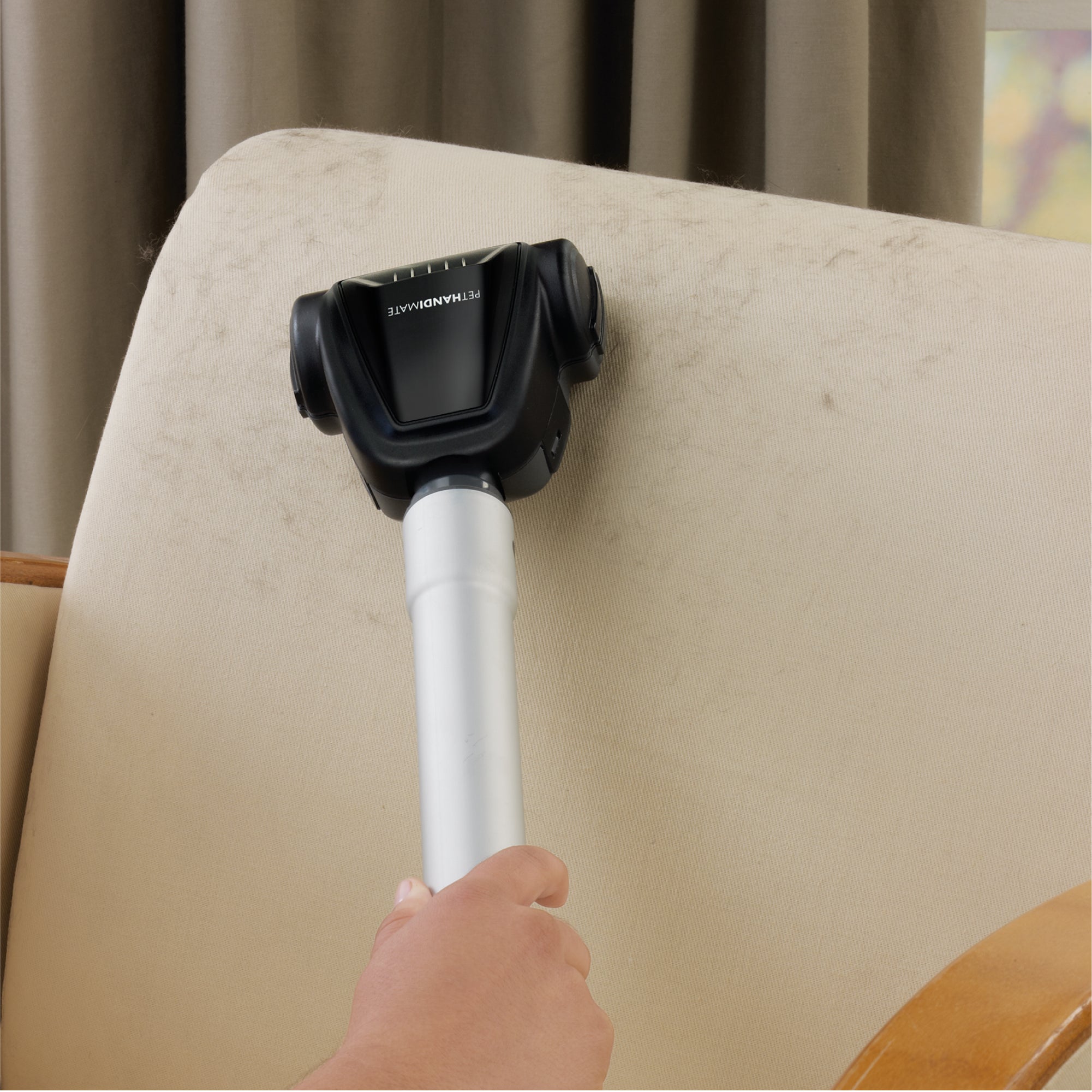 Hand using a vacuum cleaner wand and accessory tool on a beige sofa