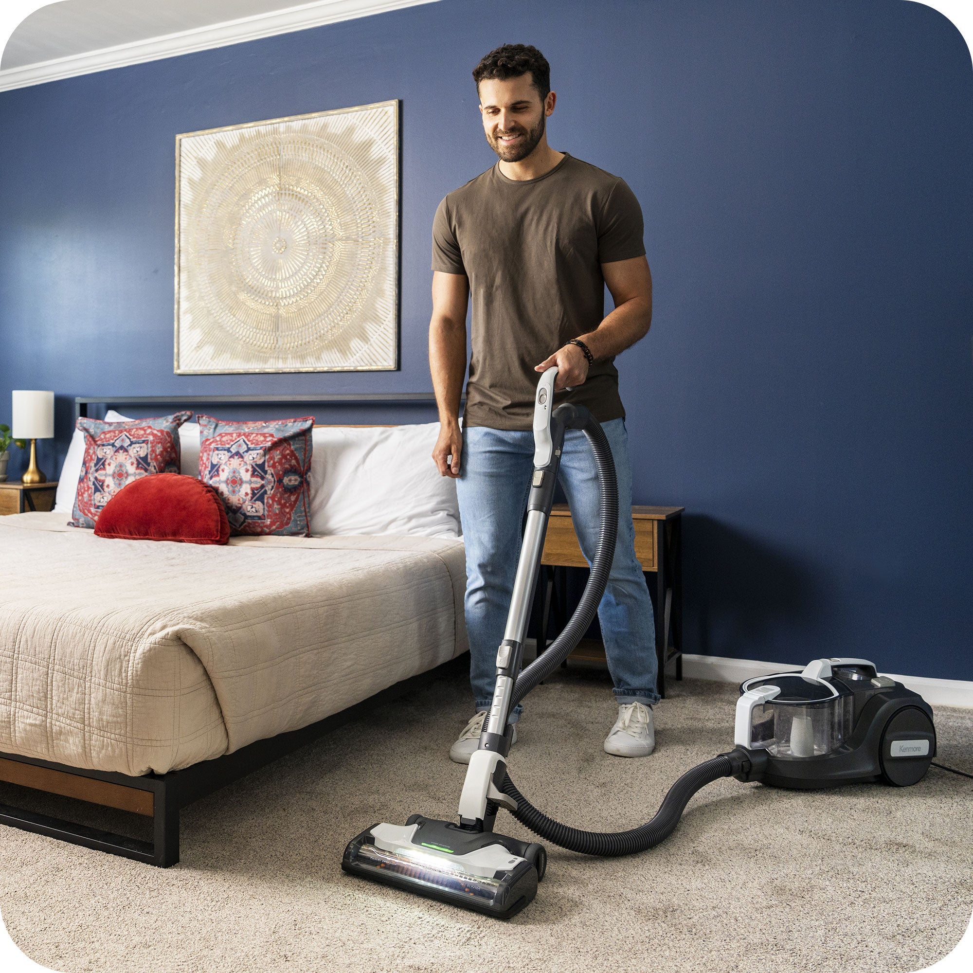 Man using a canister vacuum cleaner in a bedroom with blue walls and a bed.
