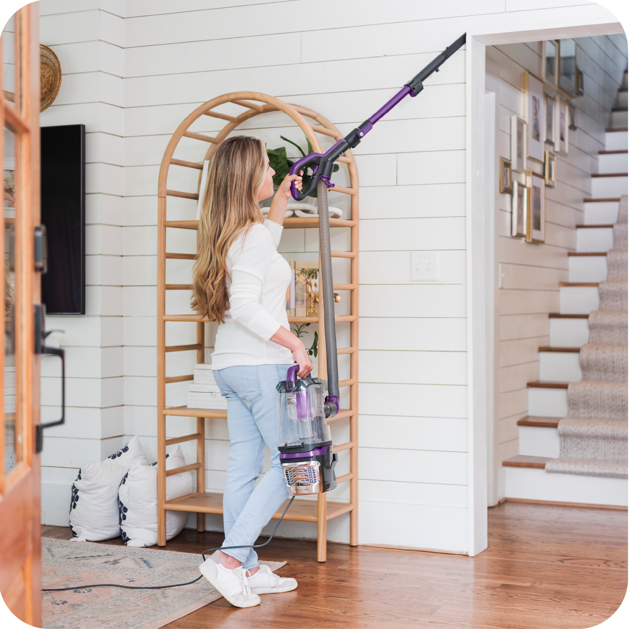Woman using a vacuum cleaner in a home setting with wooden floors and a staircase.