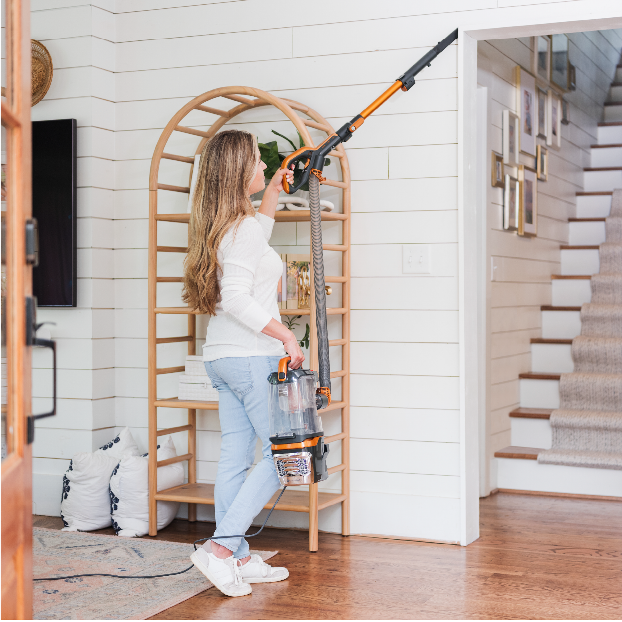 Woman using a vacuum cleaner in a home setting with wooden floors and white walls.