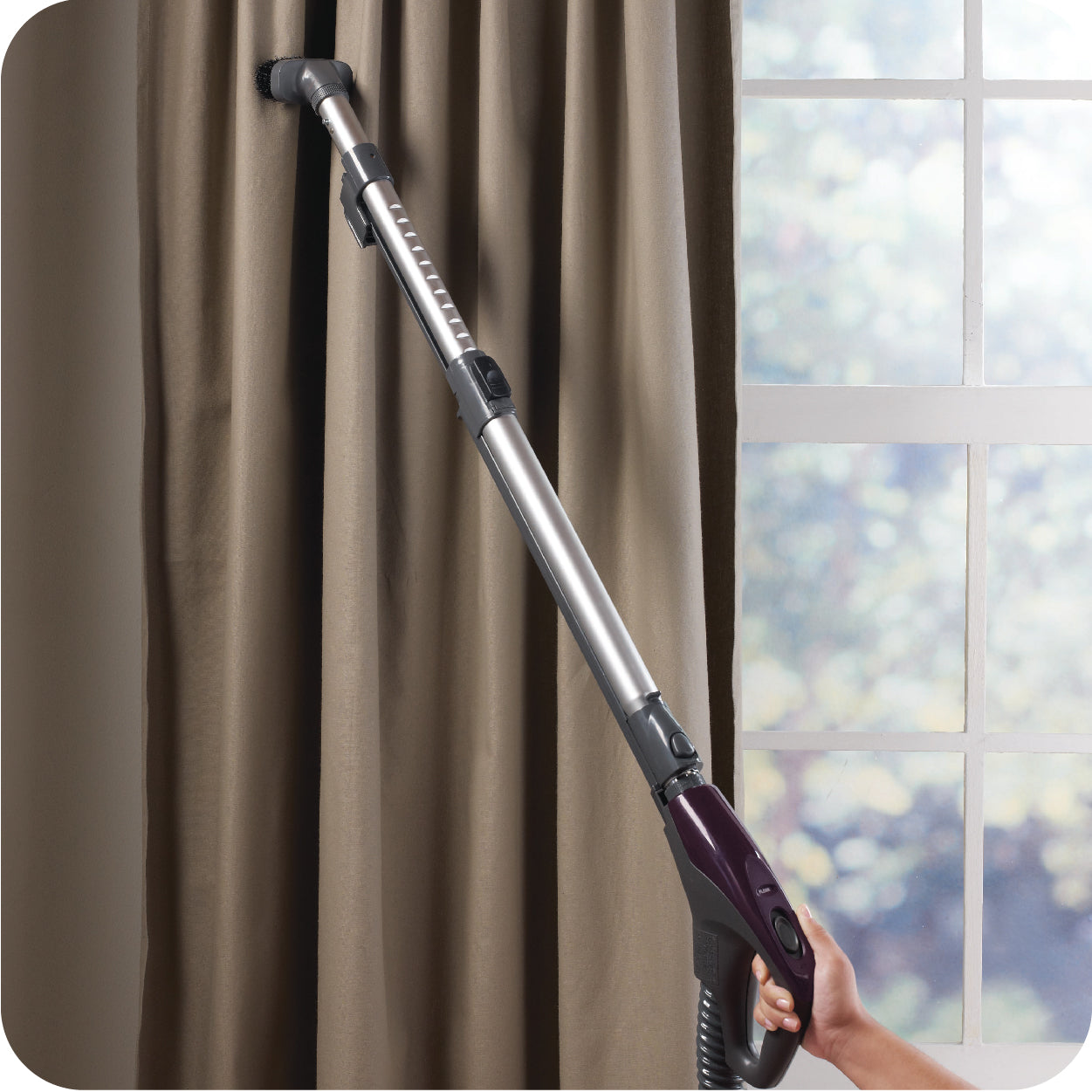 Person using a canister vacuum cleaner with a dusting brush attachment to clean a curtain near a window.