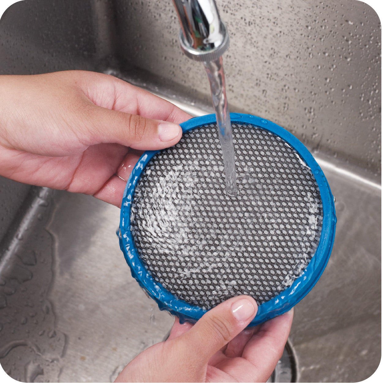 Person rinsing a vacuum cleaner filter with blue rim under running water in a sink.