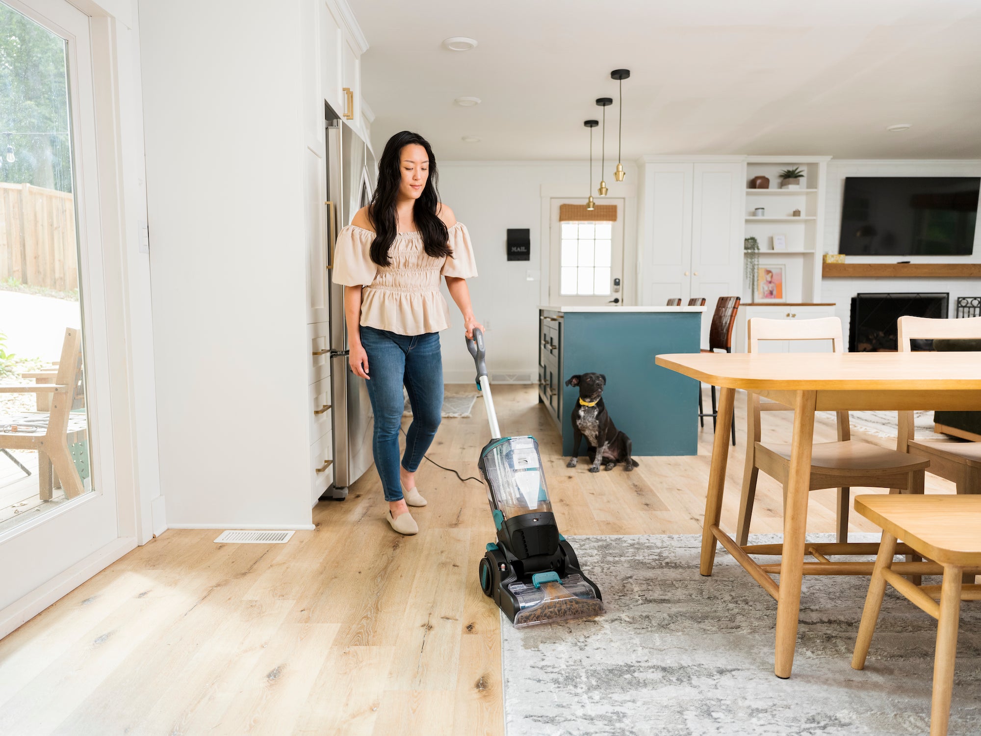Woman using a carpet cleaner in a modern kitchen with a dog and dining table.
