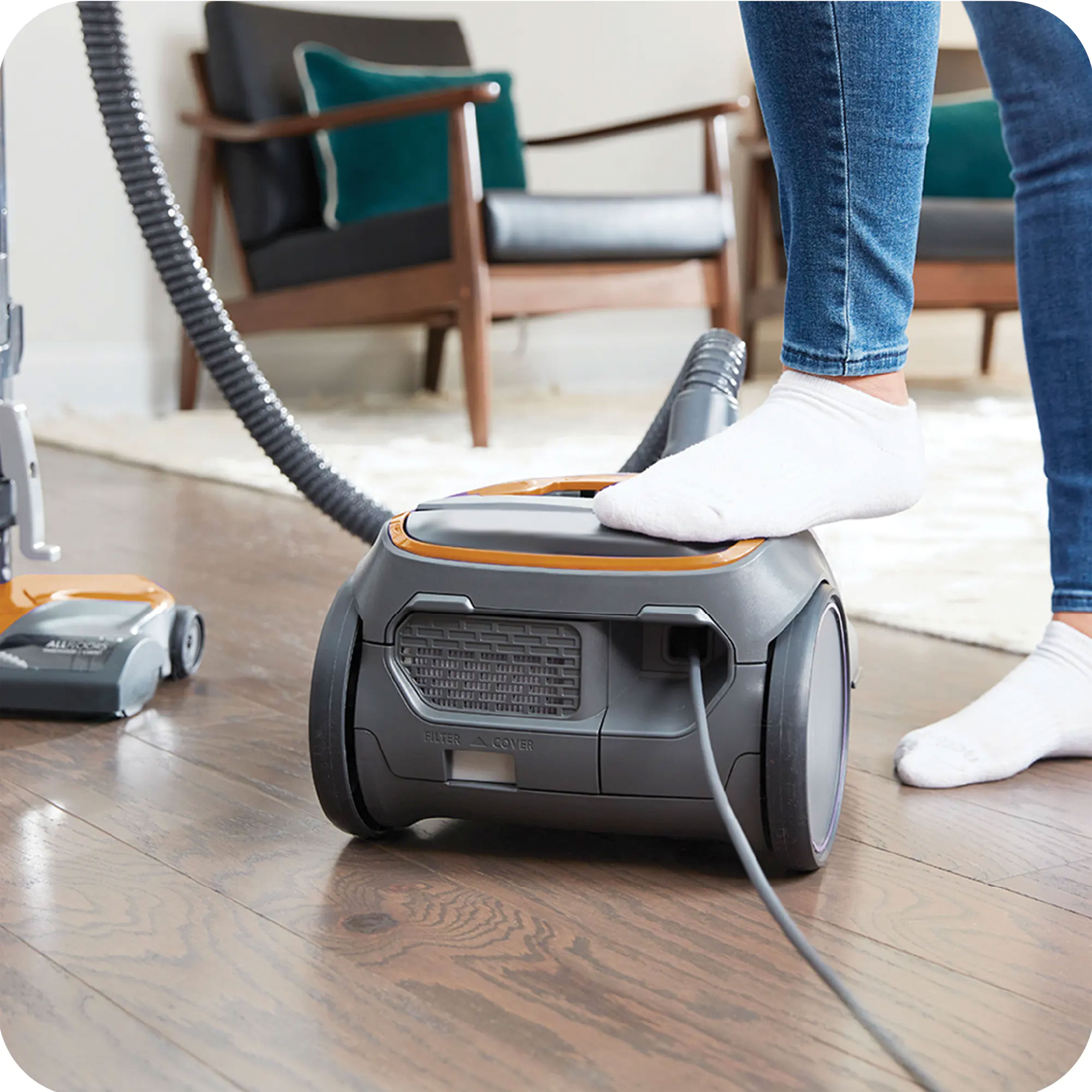 Person pressing the auto cord rewind button with their foot on the body of a canister vacuum cleaner with living room in the background