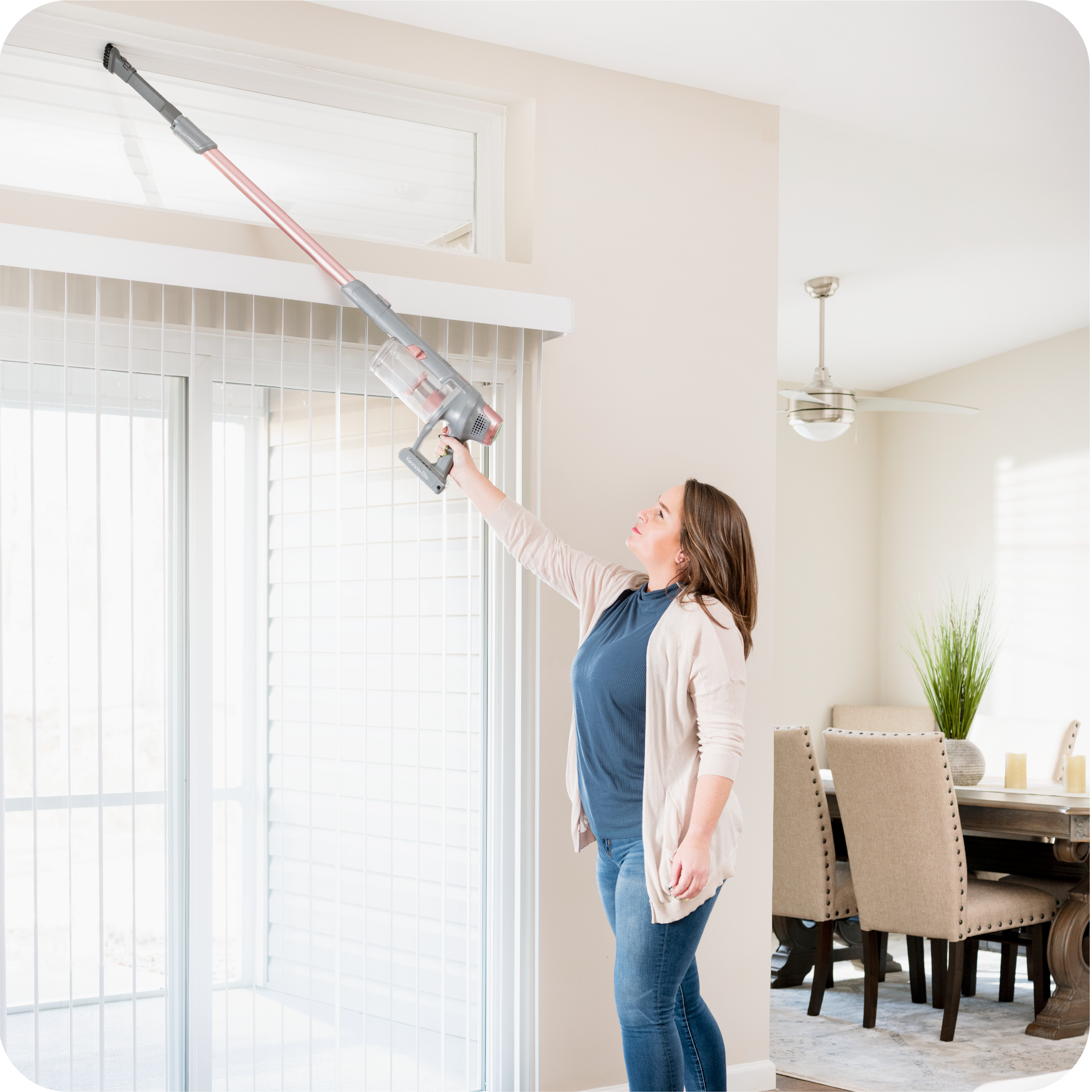 Woman using a stick vacuum cleaner to clean blinds in a room with a dining table and chairs in the background.
