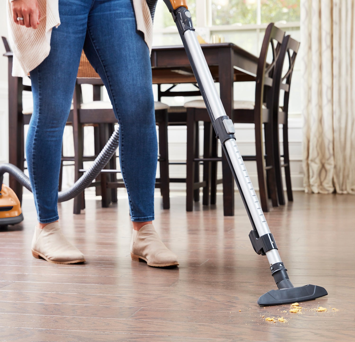 Person using a canister vacuum cleaner on a wooden floor in a room with chairs and a table.
