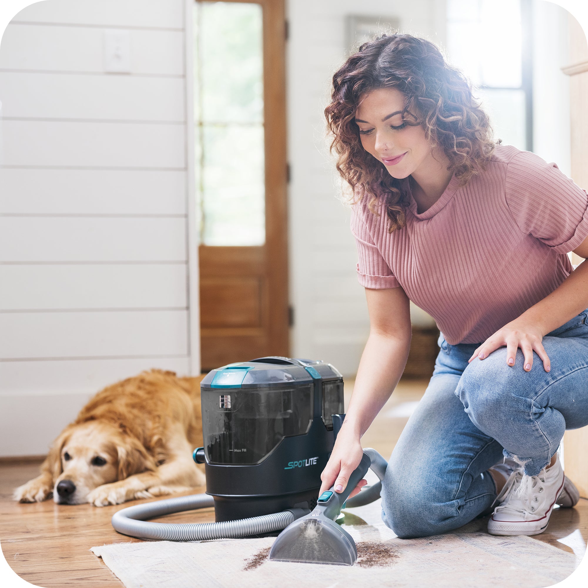 Woman knees on rug to clean dirt from carpet with a Kenmore SpotLite, dog laying in background.