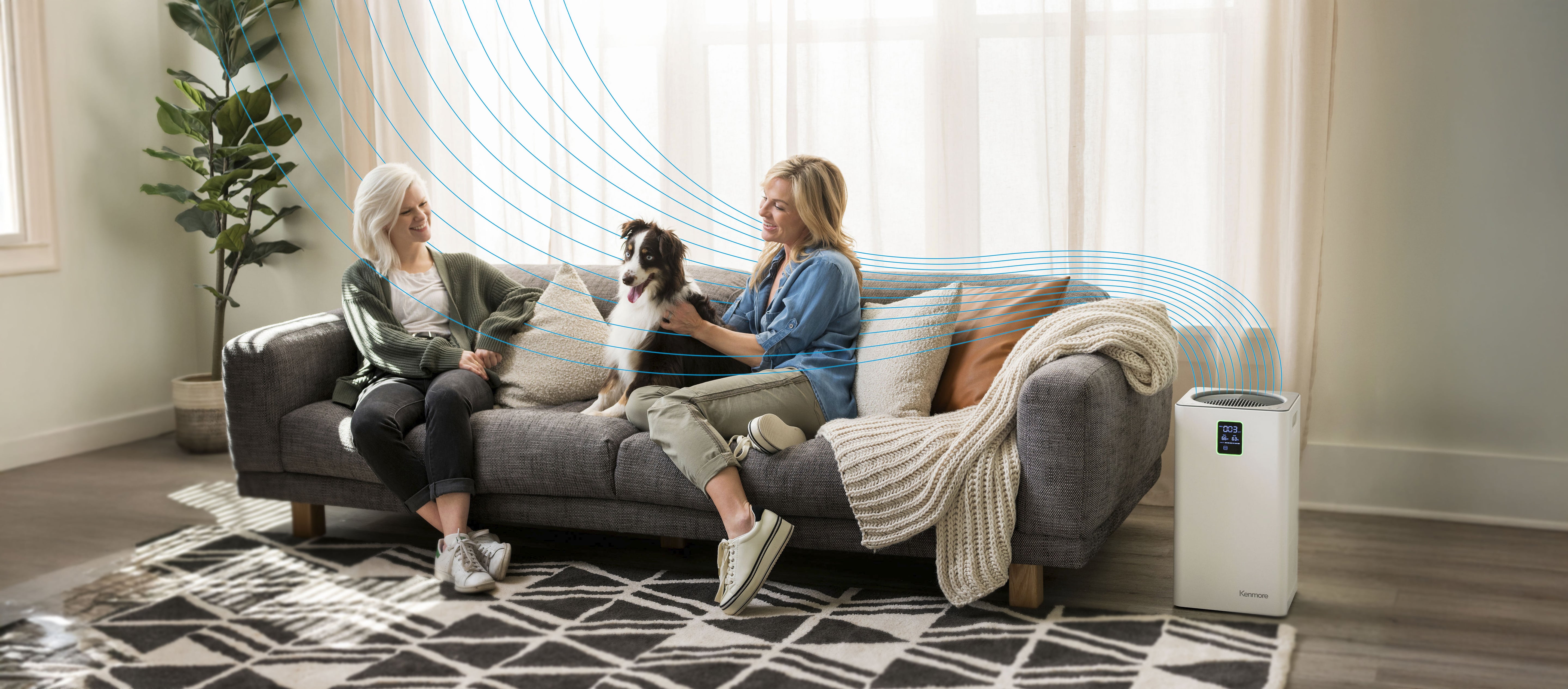 Two women sitting on a couch with a dog, an air purifier on the floor, and a plant in the background.
