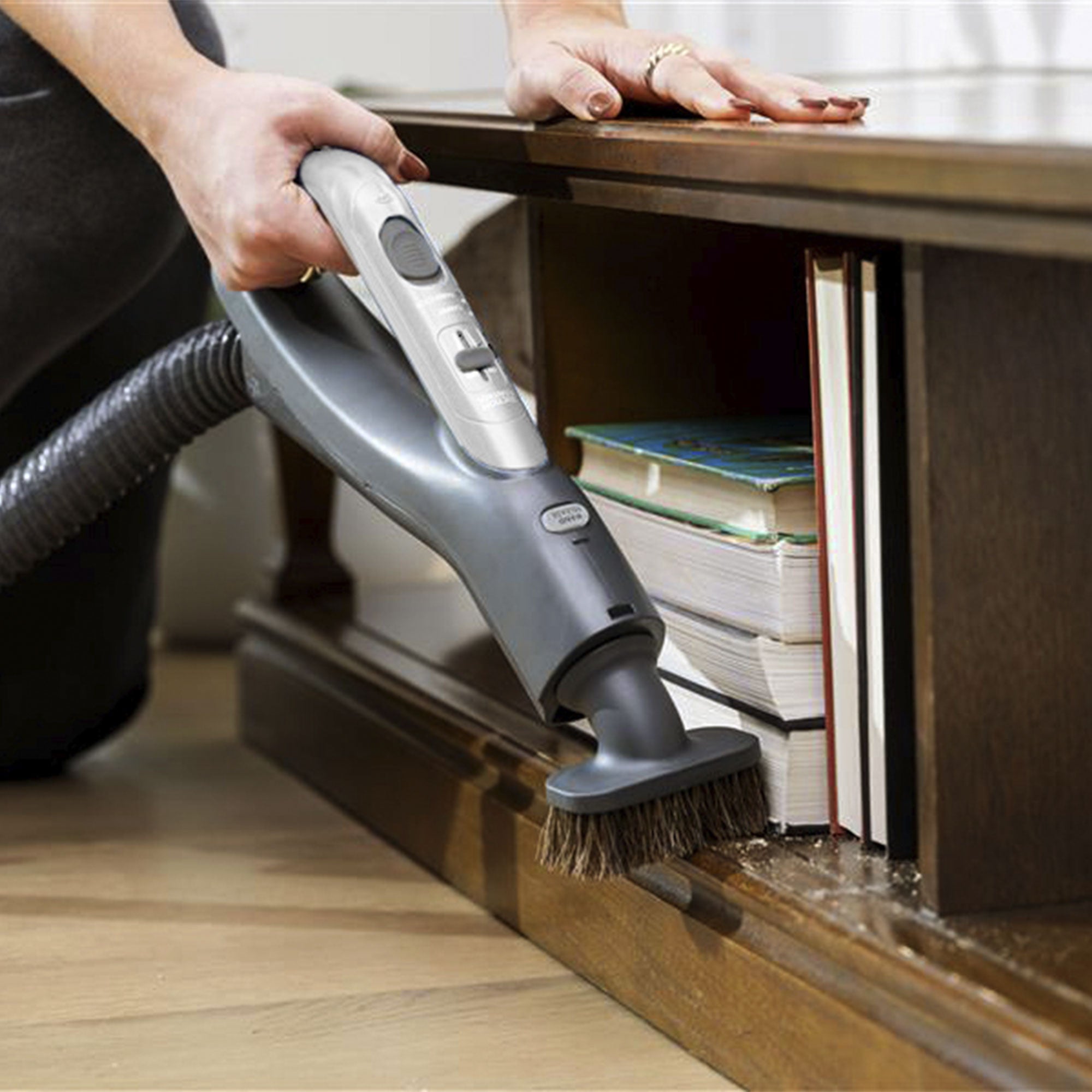 Person using a canister vacuum cleaner with a dusting brush attachment tool to clean under a bookshelf.
