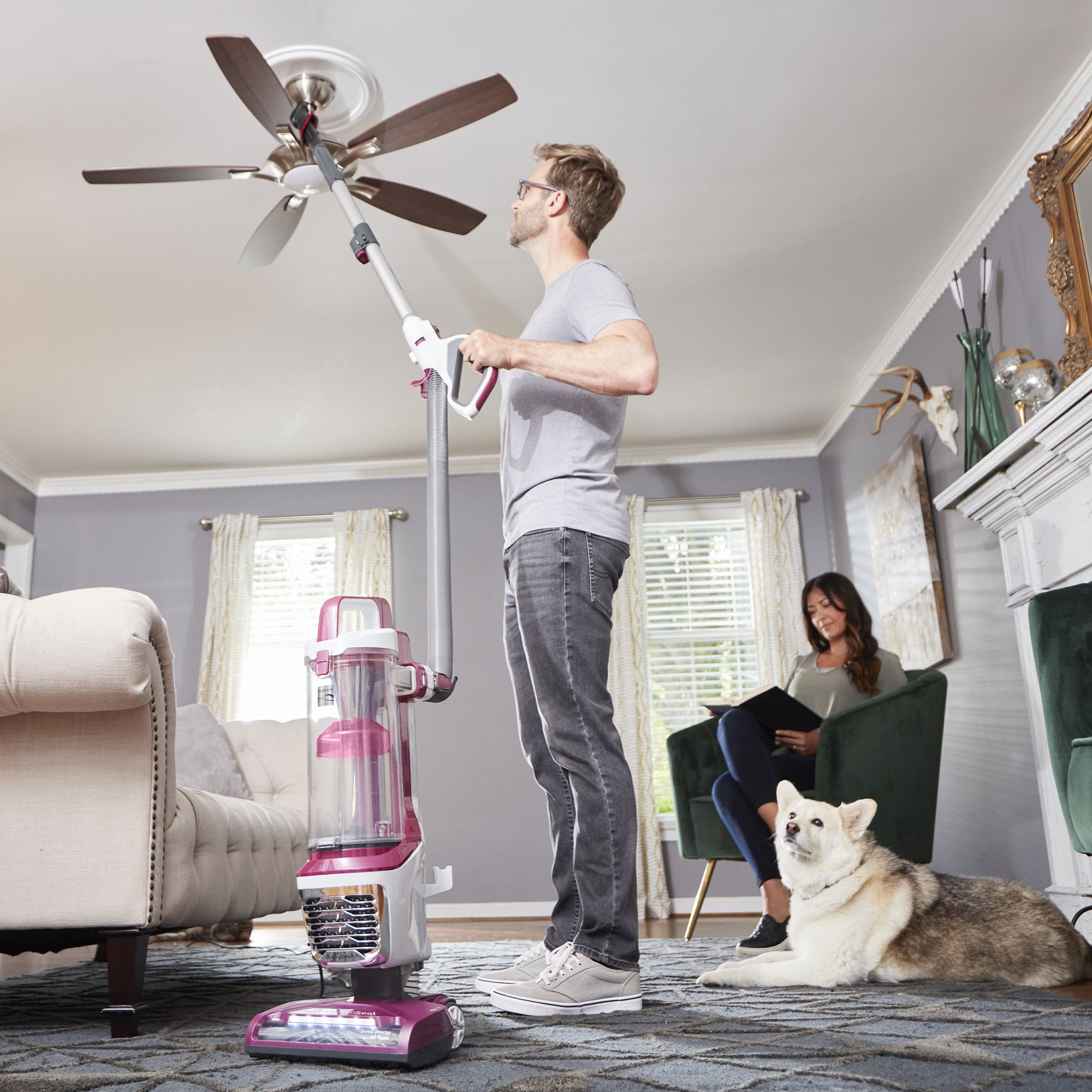Man using an upright vacuum cleaner with an attachment tool in a living room with a woman and dog.