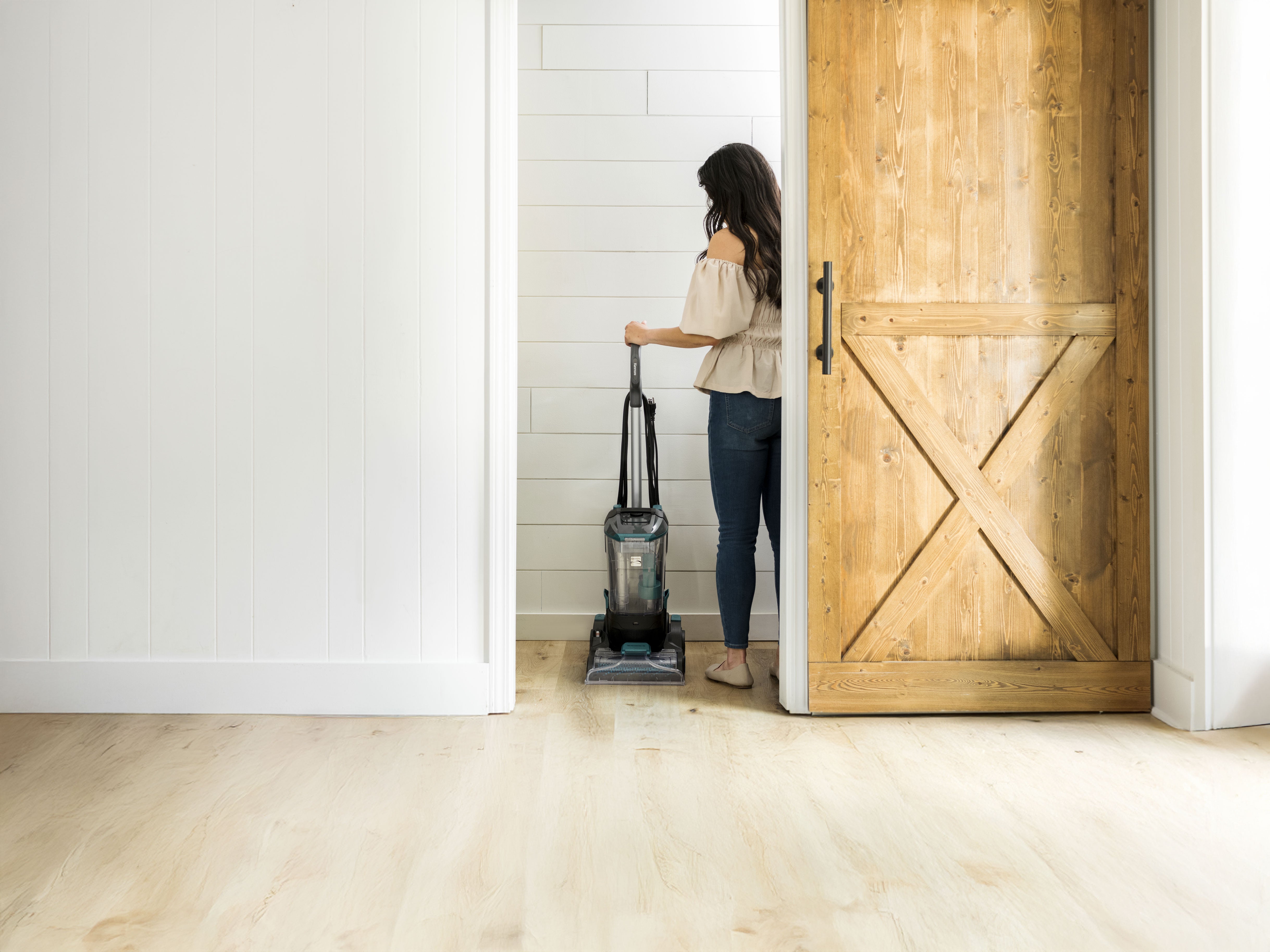 Woman storing a full-size carpet cleaner in a closet with wooden flooring and a wooden door.