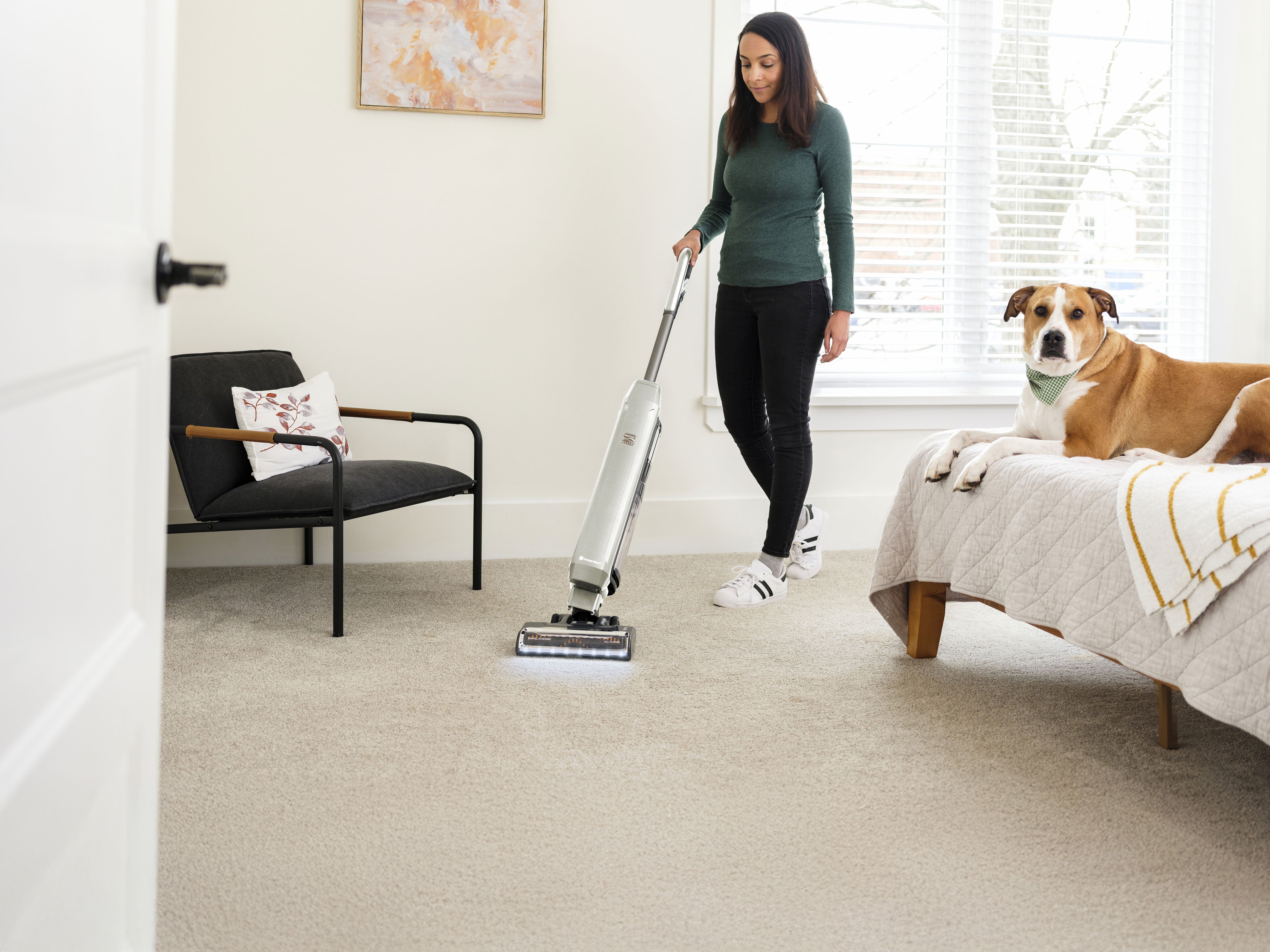 Woman using a vacuum cleaner in a living room with a dog on a bed.