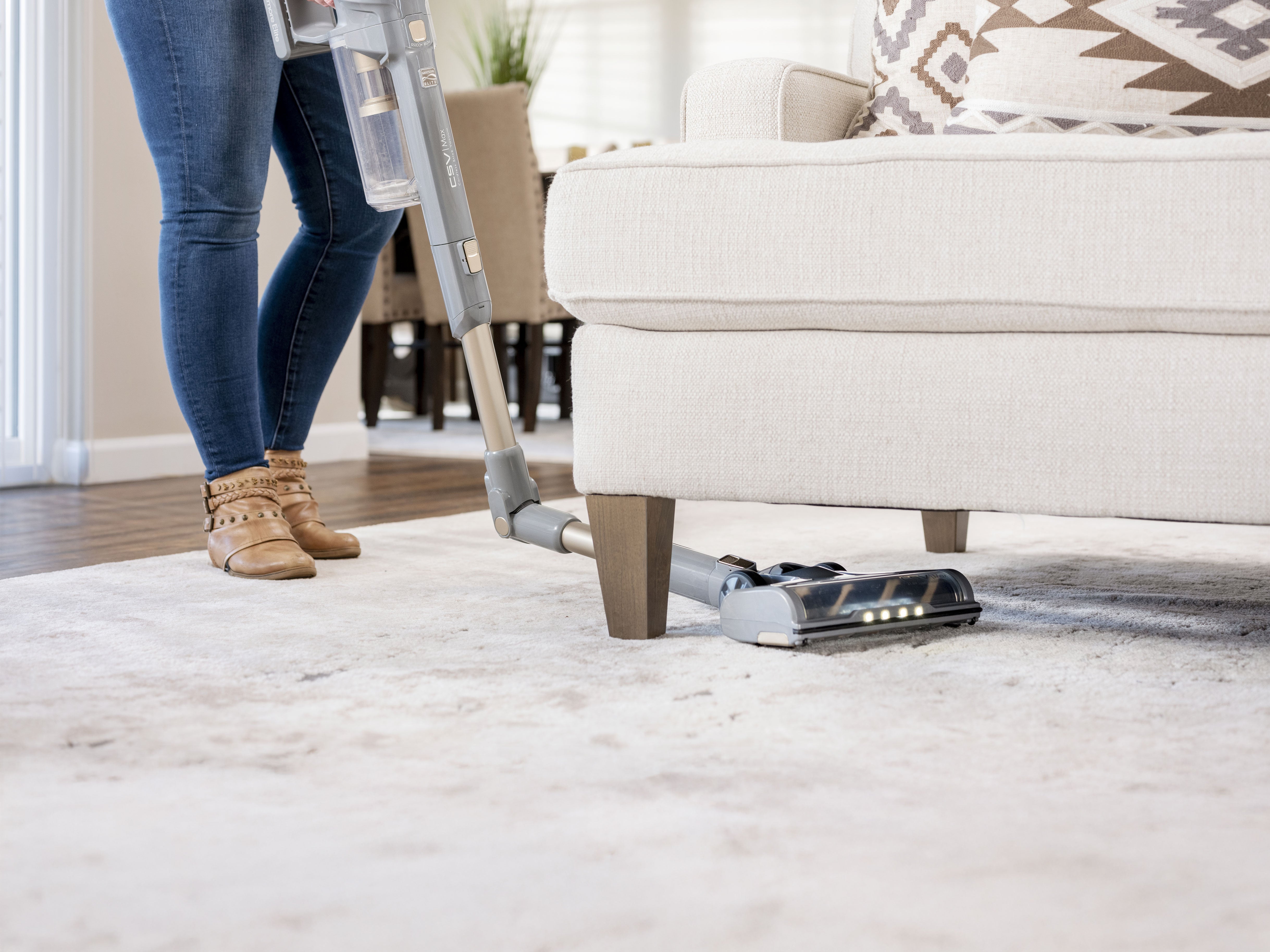 Person using a vacuum cleaner on a carpeted floor to clean underneath a couch in a living room.