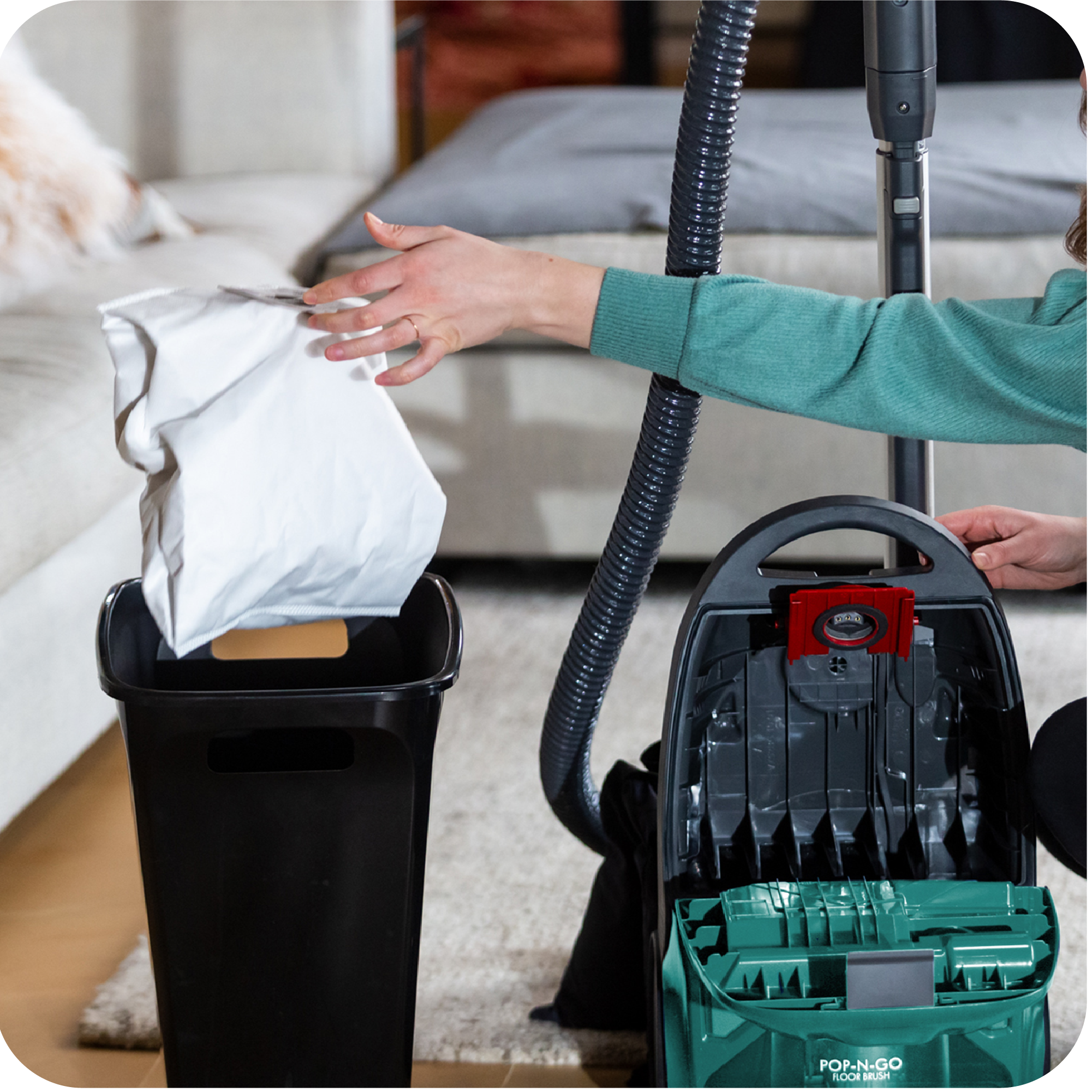 Person throwing away a used vacuum cleaner bag into a trash can in a living room.