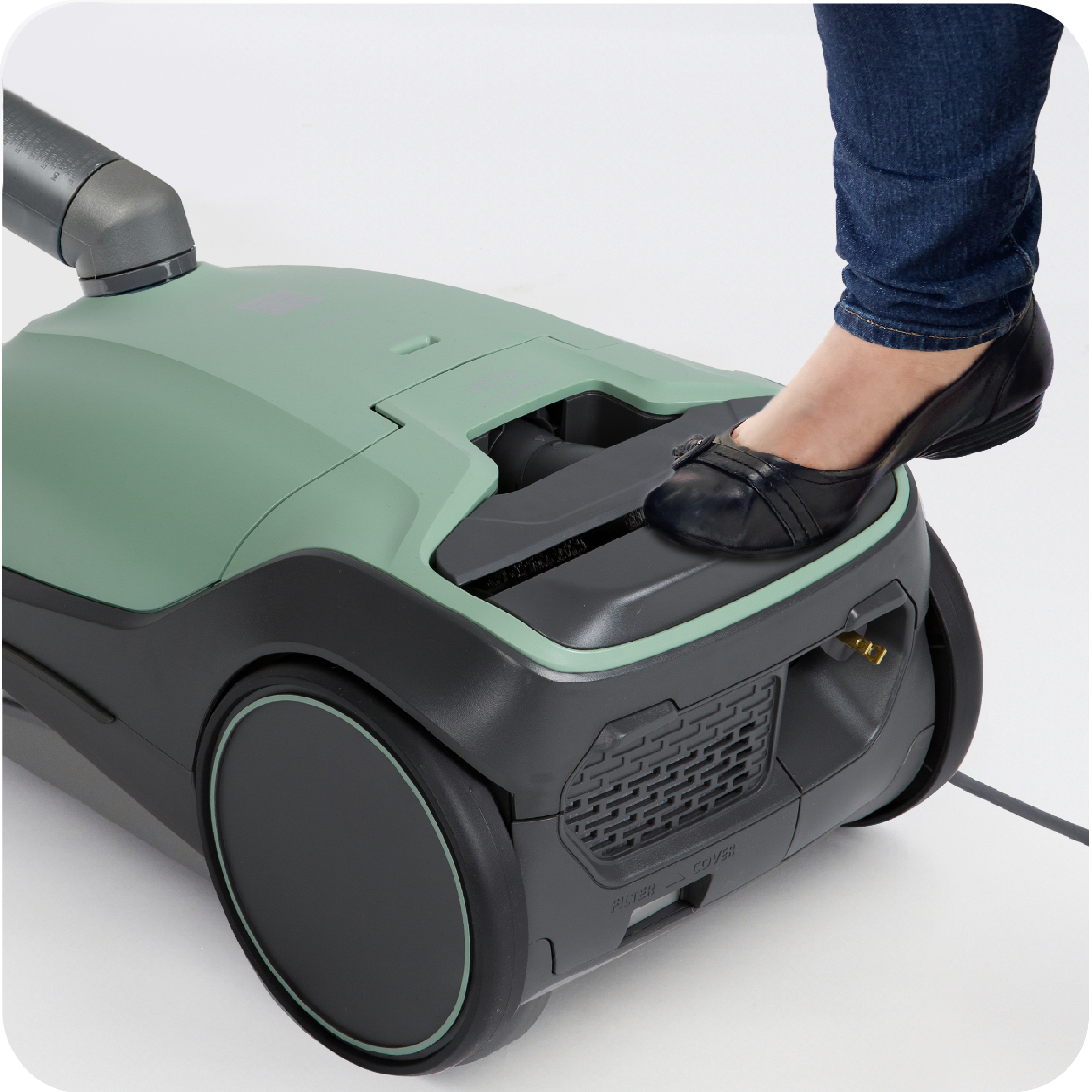 Person pressing on the auto cord rewind button with her foot on the body of a green and black canister vacuum cleaner on a white background
